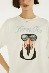 Remera blanca de algodón con cuello redondo y mangas cortas, de corte holgado. En el frente presenta el texto "Farm Rio" en tono celeste y un estampado gráfico de un tucán con anteojos de sol que reflejan un paisaje playero.
