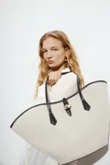 Bolso tipo shopper de tela color beige con diseño geométrico y detalles en cuero sintético negro. Tiene doble asa de mano y cierre superior con detalle metálico dorado.
