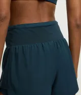 Bermuda deportiva de microfibra color azul petróleo, con diseño de doble capa que incluye un short interno ajustado y una capa externa suelta. Presenta cintura alta, bajo redondeado y bolsillo funcional en el short interno.