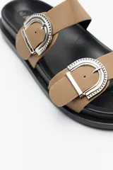 Sandalia tipo slide color tostado, con doble tira ancha y hebillas metálicas plateadas con diseño texturado. Su base es ergonómica y liviana, de color negro.