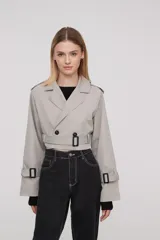 Trinchera corta color gris claro, con solapas, doble botonadura, cinturón y mangas largas con trabillas ajustables.