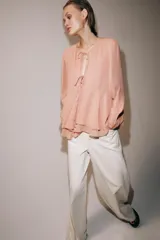 Blusa de viscosa color rosado con cuello redondo, manga larga abullonada con puños elásticos y cierre frontal con lazadas. Presenta un diseño con volantes en el bajo y corte asimétrico.