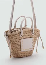 Bolso tipo cesta de fibra natural trenzada, con detalles en cuero sintético color crema. Incluye dos asas cortas de mano, una correa larga bandolera desmontable y un bolsillo frontal decorado con tachas metálicas doradas.