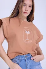 Remera color camel de algodón con mangas cortas con efecto de hombrera y estampado con una palmera y la frase "Welcome to La Guia Summer Vacation".