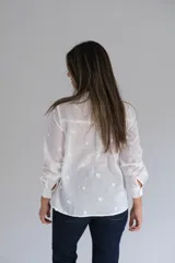 Blusa blanca de manga larga con cuello mao y cierre frontal de botones ocultos. Presenta un delicado diseño con bordados florales y detalles calados verticales en el frente.