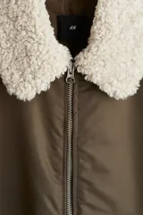 Campera bomber de nylon color marrón, ligeramente acolchada, con cuello de peluche blanco y cierre frontal. Incluye un bolsillo con cierre en una manga, bolsillos laterales con solapa y botón a presión oculto, y un bolsillo interior con botón a presión. Tiene remate acanalado ancho en puños y bajo, y un corte holgado.