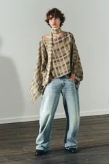 Capa corta con estampado a cuadros en tonos mostaza, marrón y blanco. Presenta un cuello redondo y mangas amplias tipo murciélago. Incluye un lazo o foulard integrado en el cuello para anudar.