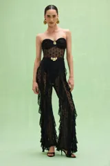 Mono strapless negro confeccionado en encaje con diseño calado. Presenta un corpiño con detalle de frunce y hebilla dorada en el centro, cintura marcada con cinturón fino y hebilla dorada, y piernas acampanadas con volados laterales de encaje.