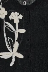 Chaqueta de punto negra con cuello redondo y manga larga. Presenta bordados florales en color blanco en la parte delantera y cierre frontal con botones.