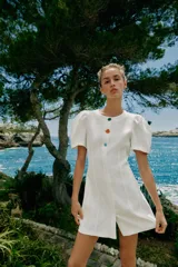 Vestido corto de color blanco con cuello redondo y mangas cortas abullonadas. Presenta un cierre frontal con botones decorativos de colores.