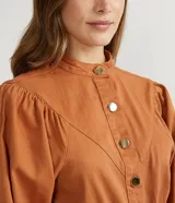 Mono corto color ladrillo, de sarga de algodón, con cuello mao, mangas abullonadas largas, cierre frontal con botones dorados, bolsillos laterales y cintura marcada.