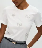 Remera de algodón de corte regular, con cuello redondo y mangas cortas. Presenta aplicaciones de lazos decorativos realizados con pedrería brillante en la parte frontal.
