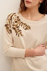 Sweater color beige de punto suave, con cuello redondo, manga larga y diseño holgado. Presenta un delicado bordado de ramas y hojas con cuentas metalizadas en la parte superior derecha del pecho y el hombro.