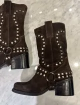 Bota de caña media confeccionada en cuero gamuzado color marrón, con diseño de estilo western. Presenta detalles de tachas metálicas plateadas distribuidas en la caña y en una correa desmontable con argolla metálica en el tobillo. Cuenta con taco cuadrado y suela de goma.