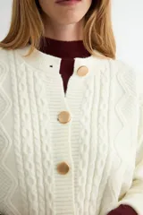 Cardigan de punto trenzado color crema con cuello redondo y cierre frontal de botones grandes en tono dorado. Presenta mangas largas con puños acanalados y un diseño de tejido con textura.