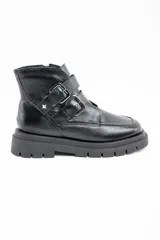 Bota corta de cuero negro con suela track dentada y dos correas decorativas con hebillas metálicas en el lateral. Presenta un diseño de puntera cuadrada y cierre lateral.
