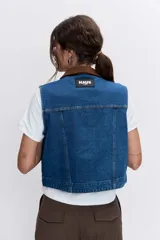 Chaleco de denim azul con cuello camisero en contraste color marrón, cierre frontal con botones metálicos y bolsillos superiores con solapa.