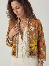Chaqueta corta de terciopelo color camel con bordados coloridos de inspiración folclórica que incluyen pájaros blancos, corazones rosados, estrellas amarillas y figuras, con cuello redondo y diseño abierto al frente.