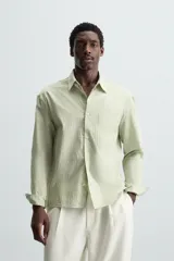 Camisa de hombre de corte relajado (relaxed fit) en color verde menta pálido, confeccionada en tejido con textura irregular acanalada. Presenta cuello solapa, manga larga con puño abotonado, bolsillo plastrón en el pecho y bajo asimétrico con aberturas laterales.