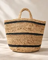 Bolso de mimbre tejido en color arena con detalles de líneas horizontales negras y dos asas cortas superiores.