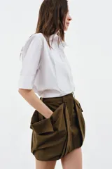 Camisa de algodón con cuello solapa y mangas por debajo del codo. Presenta un diseño con pechera plisada frontal, bajo recto y cierre mediante botones ocultos bajo solapa.