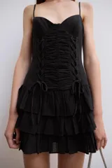 Vestido corto negro de tirantes finos ajustables y escote en V. Presenta un diseño ajustado con drapeado central y cordones cruzados laterales tipo corsé, y falda corta con tres capas de volantes escalonados.