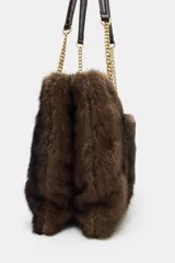 Bolso de hombro con cuerpo de textura efecto pelo en color marrón oscuro. Cuenta con un asa de hombro combinada con cadena metálica dorada, bolsillo exterior frontal y cierre principal mediante cremallera.