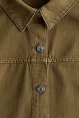 Campera camisera de sarga de algodón color verde militar, con cuello clásico, cierre frontal de botones metálicos, canesú y bolsillos de parche delanteros.