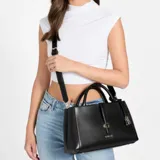 Cartera Guess modelo Carrie de color negro, confeccionada en símil cuero con herrajes dorados. Presenta doble asa superior, una correa ajustable para el hombro y un dije decorativo con candado. Cuenta con el logo de la marca en el frente y una tira con cierre de broche magnético.