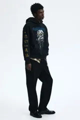 Canguro negro de corte oversized con capucha, bolsillo frontal tipo canguro y estampado frontal de la banda Iron Maiden con diseño de momia. Las mangas presentan detalles estampados en color dorado con motivos egipcios.