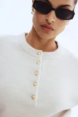 Buzo de punto suave color beige claro, con cuello redondo y abertura frontal con botones dorados. Presenta hombros caídos y puños y bajo acanalados.