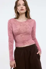 Remera rosa de manga larga, confeccionada en tela con transparencias y detalles en flock con estampado paisley. Tiene cuello a la base, corte al cuerpo y frunces laterales.