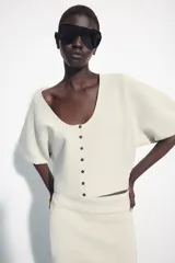 Chaqueta corta de punto color blanco crudo, con escote redondo pronunciado y cierre frontal con botones oscuros. Presenta mangas cortas abullonadas con frunces.