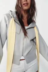 Chaqueta de punto con capucha, manga larga y cierre frontal con cremallera. Presenta un diseño de lazo decorativo en color amarillo en la zona del cuello.