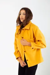 Camisa color mostaza de paño, corte oversize, con cuello clásico, cierre frontal con botones, mangas largas con puños abotonados y dos bolsillos de parche en el pecho.