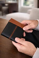 Billetera de cuero para hombre con diseño bicolor, combinando tonos marrón y negro. Cuenta con solapa central, múltiples ranuras para tarjetas, compartimento para billetes y visor transparente. Presenta el logo de la marca grabado en el frente.