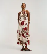 Vestido midi de corte evasé, con escote halter y abertura frontal en el busto, anudado en la parte superior. Presenta un estampado grande de flores y hojas en tonos rojo oscuro, beige y negro sobre fondo crudo. El diseño incluye un volante fruncido en el ruedo inferior.