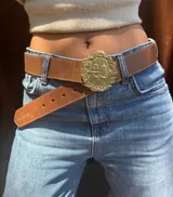 Cinturón de cuero marrón con hebilla metálica grande en forma de escudo nacional.