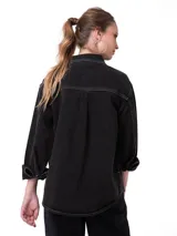 Camisa oversize de denim negro con costuras en contraste color blanco, bolsillos dobles en el pecho con solapa, cierre frontal de botones metálicos y dobladillo redondeado.