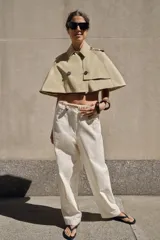 Trench corto tipo capa de algodón en color beige, con cuello solapa, trabillas en los hombros, bajo redondeado y cierre frontal cruzado con botones.