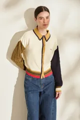 Cardigan de punto 100% algodón, diseño color block con cuerpo blanco, manga derecha verde militar y manga izquierda azul marino, ambas con tejido trenzado. Cuenta con cuello polo mostaza con ribete oscuro, cierre frontal con botones y detalles de rayas en el elástico inferior en tonos mostaza, marrón y fucsia.