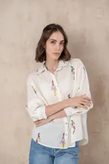 Camisa blanca de manga larga con cuello clásico y cierre con botones. Presenta un estampado bordado con figuras humanas abstractas en colores.