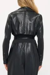 Trench largo de cuero sintético negro con solapas anchas, cierre frontal cruzado y cinturón ajustable en la cintura. Presenta detalles de tejido acanalado en la zona de la cintura y bolsillos laterales.