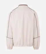 Campera de tejido técnico color beige con detalles de costuras en contraste color marrón. Presenta cuello alto, cierre frontal con cremallera, mangas largas con puños elásticos y estampa frontal con texto. Incluye un detalle bordado en la manga.