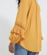 Blusa de manga larga color mostaza con textura de puntos en relieve (dobby). Presenta cuello redondo con abertura en V y cordón ajustable con borla (tassel) en el escote. Las mangas tienen detalles de entretejido calado (entremedios) y volados con terminación fruncida en los puños.
