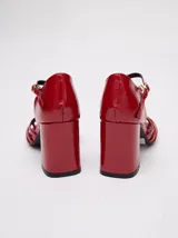 Sandalias de taco alto ancho con diseño retro, confeccionadas en charol negro. Presentan tiras delanteras múltiples entrelazadas, talón cerrado y una correa fina ajustable al tobillo con hebilla metálica.