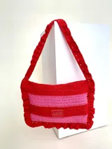 Cartera tipo shoulder bag tejida a crochet en hilo de algodón, con diseño a rayas en tonos de rojo y rosado, y detalles de volados en los bordes y la manija de hombro.