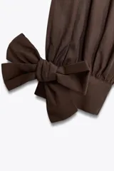 Camisa marrón de manga larga con cuello solapa y cierre frontal de botones. Los puños terminan con lazos anudados.