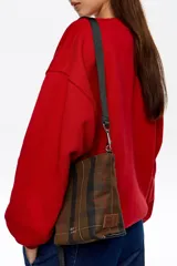 Bolso bandolera de gabardina con estampado de cuadros en tonos marrones y verdes, con asa de cinta negra regulable y logo de la marca estampado en la parte inferior.