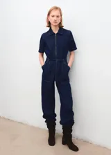 Mono de denim azul oscuro con cierre frontal de cremallera, cuello camisero y mangas cortas. Presenta un corte recto, bolsillos de parche delanteros y traseros, y un cinturón de la misma tela para ajustar la cintura.